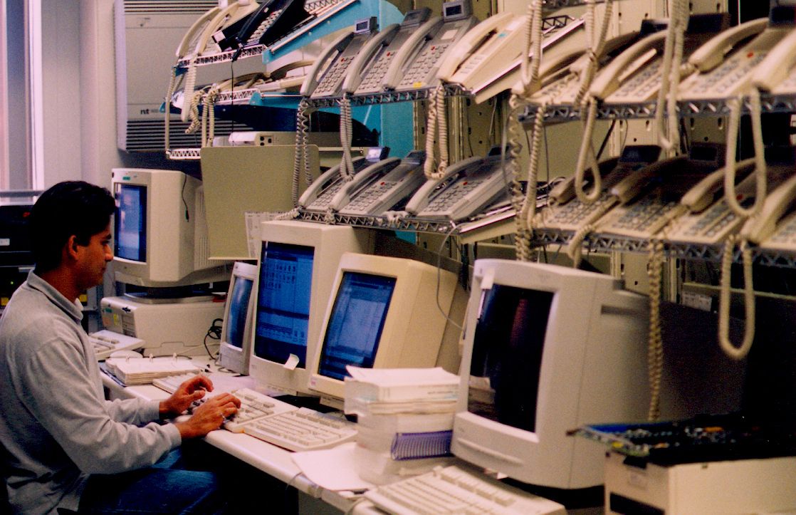 A man sits at a desk with five computer stations underneath shelves holding 21 telephones at Nortel, Belleville, Ontario.